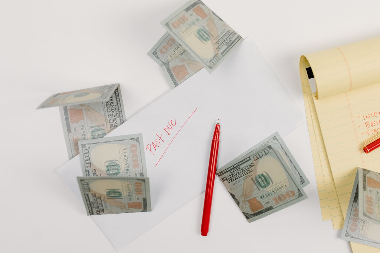 A top-down view of scattered US dollar bills with a past due envelope, red pen, and notepad.